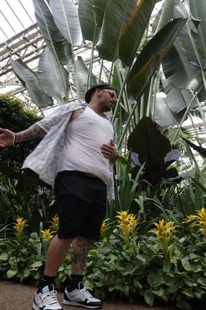 White-shirted figure posing among large banana leaves and yellow flowers in a greenhouse setting.