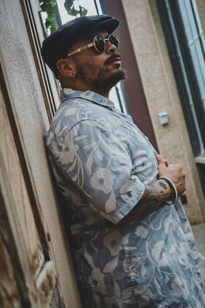 Person wearing camo jacket and baseball cap leaning against building wall in urban setting.