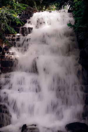 Misty white waterfall cascading down dark rocky cliffs in a lush forest setting.