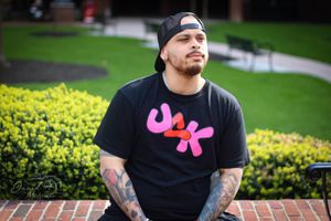 Person in black clothing with visible tattoos stands amid green garden landscaping.