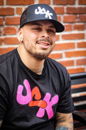 Person wearing black t-shirt with pink logo design and baseball cap poses against brick wall.
