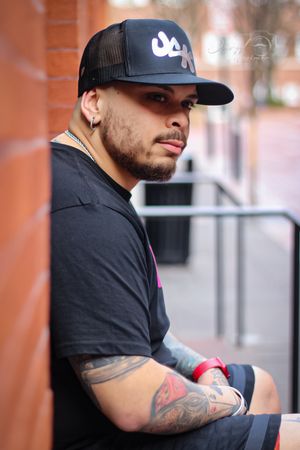 Profile view of person in black t-shirt and cap showing tattooed arm against brick background.