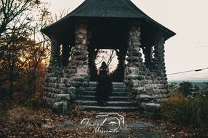 A cloaked figure poses dramatically in a stone gazebo at sunset.