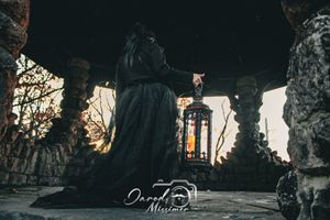 Hooded figure holding lantern in gothic stone structure at sunset.