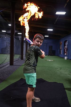A baseball player swings a flaming bat in an indoor training facility with artificial turf and netting.