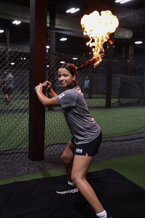 A softball player practices batting technique in an indoor batting cage with turf and netting.