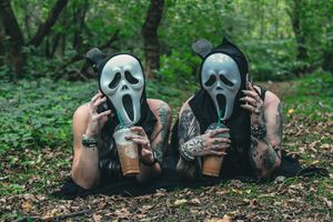 Two people lying in grass wearing spooky ghost masks and dark clothing.