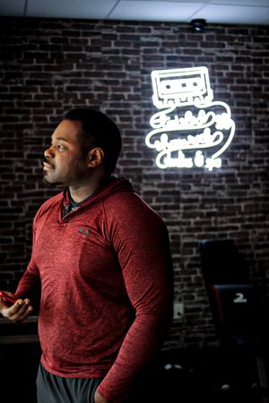 Red-shirted person standing against brick wall with neon sign.
