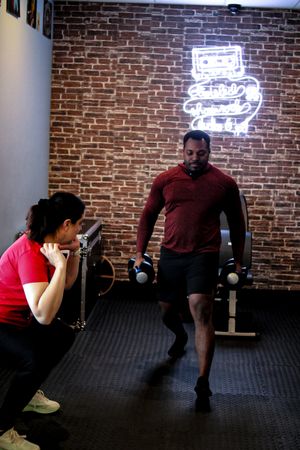 Two people working out in a gym with weight training equipment and exposed brick wall with neon signage.