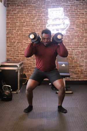 Person performing squat exercise with dumbbells in front of neon sign on brick wall.