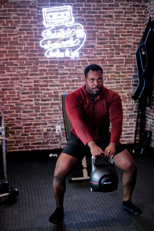 Person exercising with kettlebell in gym setting with exposed brick wall and neon sign.