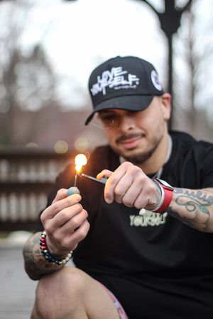 Person with tattoos lighting something while wearing dark clothing and baseball cap.