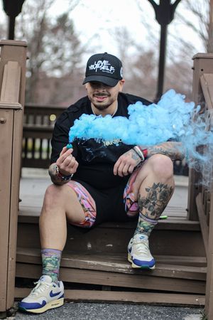 Person exhaling blue smoke while sitting on wooden deck wearing sneakers and shorts.