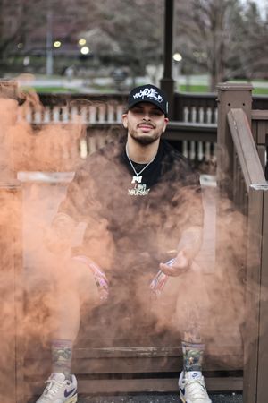 Brown smoke effects surround person sitting on wooden railing in casual streetwear.