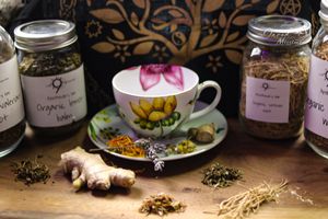 Herbal tea arrangement with decorative teacup and mason jars of dried herbs on wooden surface.