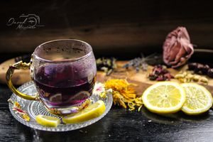 Purple tea in glass cup with lemon slices and dried rose on dark wooden surface.
