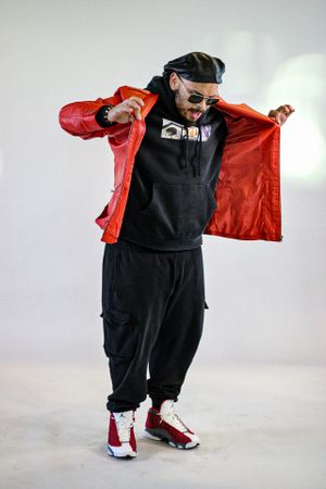 Person in baggy black clothes and red jacket performing dance moves against white background.