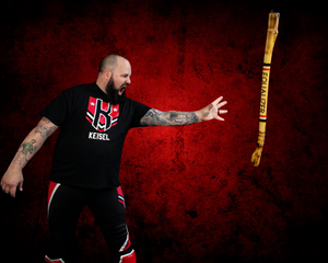 A tattooed athlete in black athletic wear reaching towards a wooden staff against a dramatic red background.