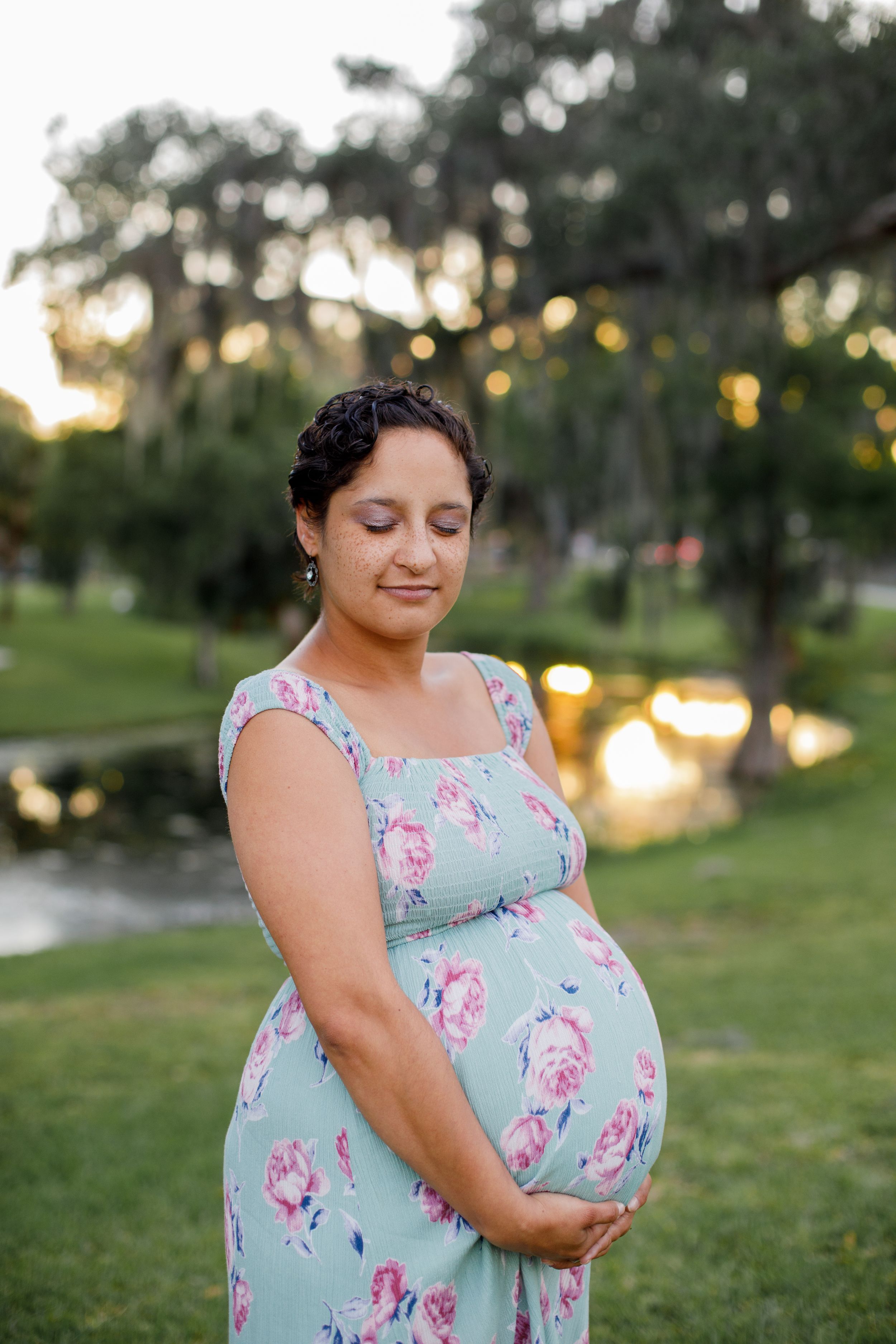Expectant mother in a mint floral dress poses in a park during sunset.