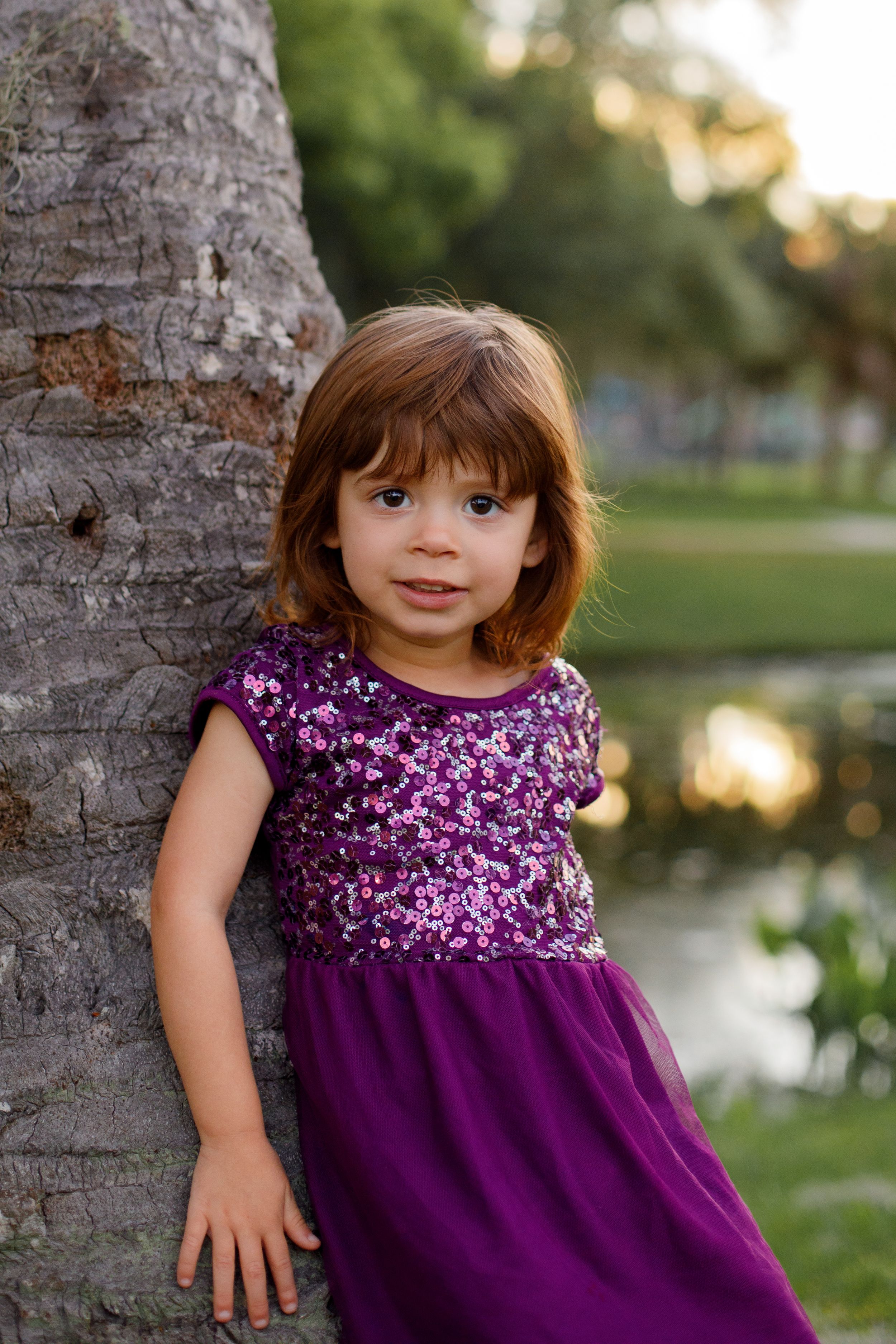 Child in purple sequined dress poses by palm trees at sunset.