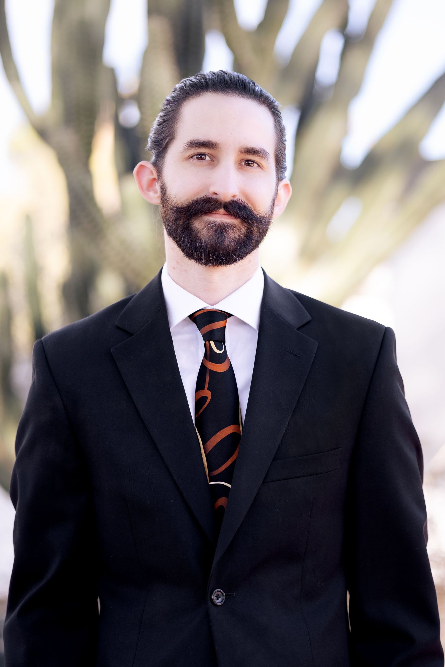 A man in a dark suit and striped tie smiles professionally against a blurred outdoor background.
