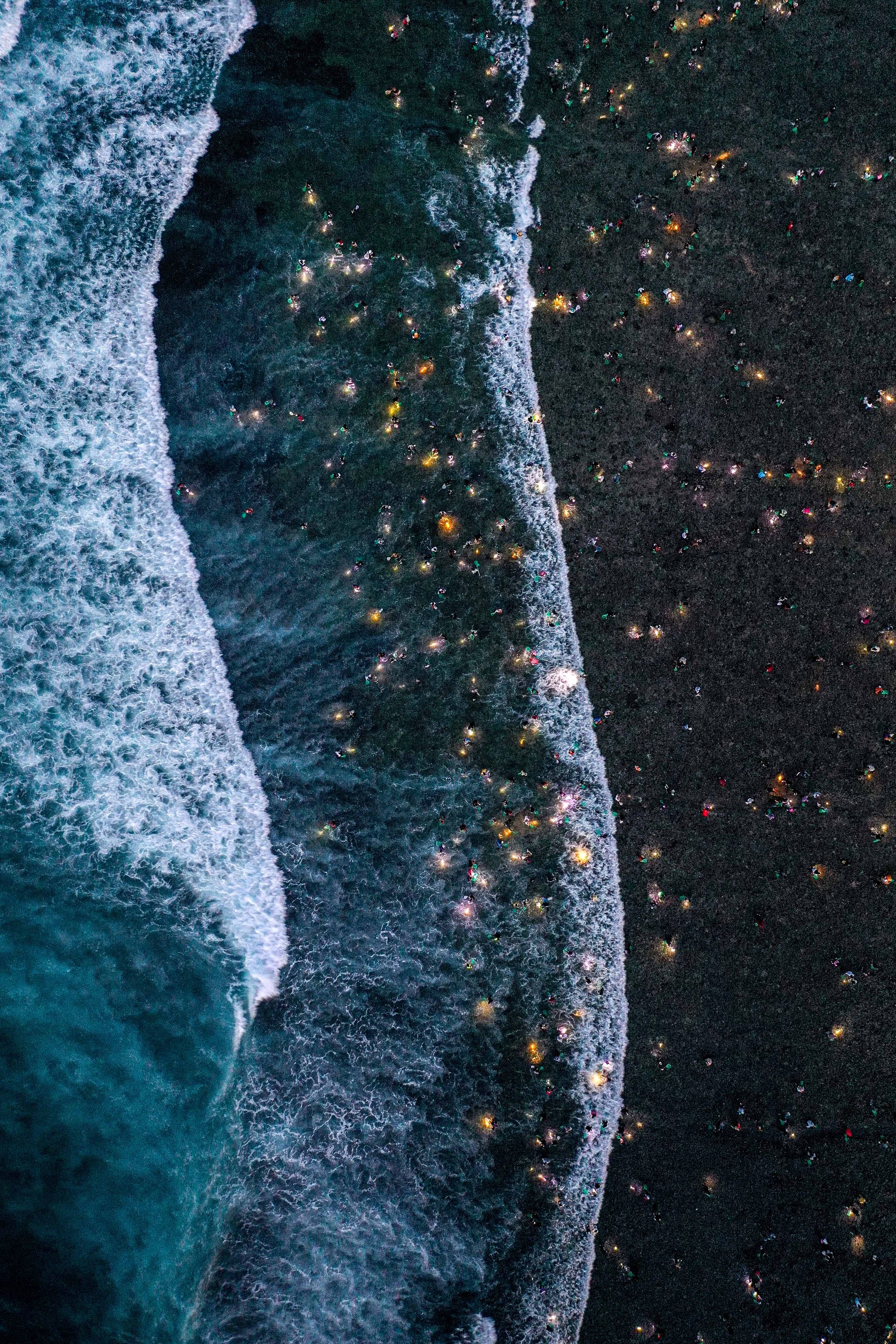 Aerial view of ocean waves meeting dark coastline with glowing city lights at night.