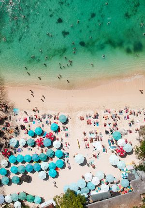 Aerial view of crowded beach with blue umbrellas and swimmers in crystal clear turquoise water.
