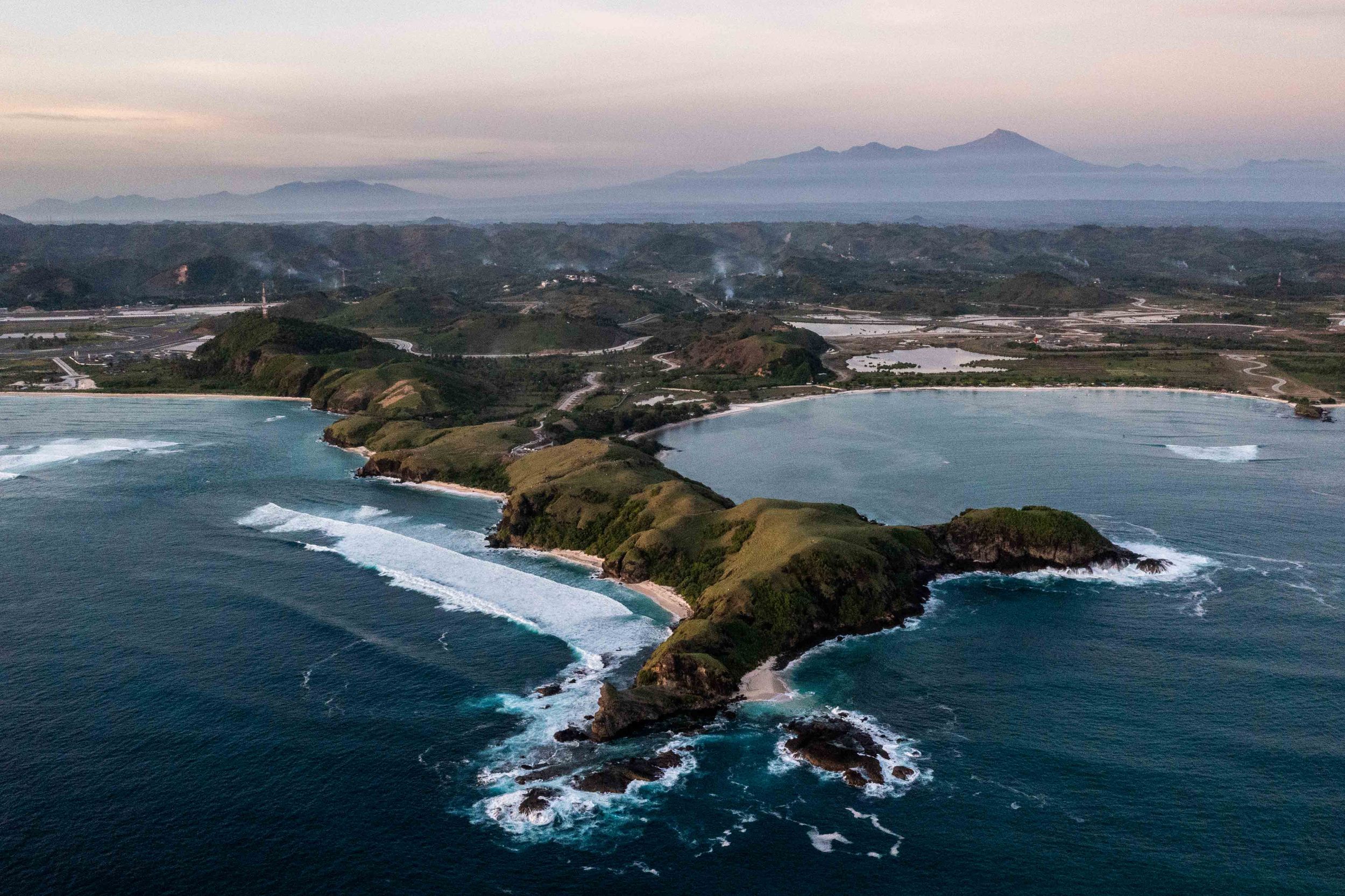 Aerial view of rugged coastline with waves crashing against cliffs and distant mountains.