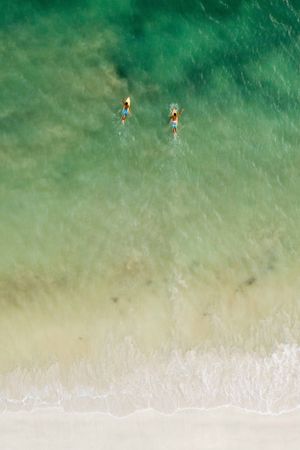 Birds-eye view of two swimmers floating in crystal clear turquoise ocean waters near a white sandy beach.