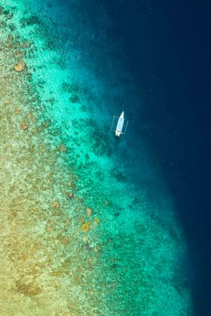 Aerial view of a lone sailboat navigating between vibrant turquoise coral reef and deep blue ocean waters.