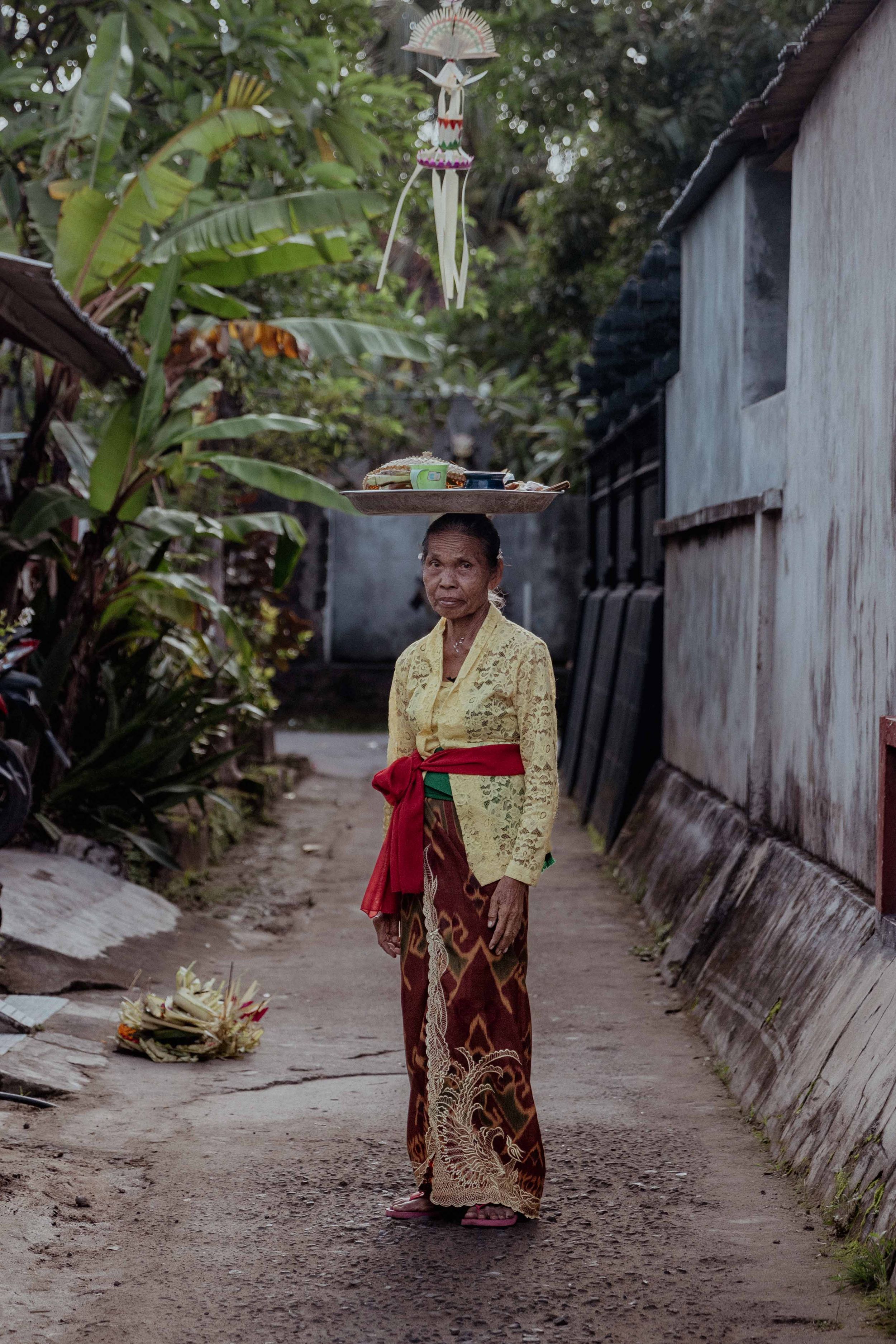 Person wearing traditional Balinese dress standing in a narrow alley with tropical plants.