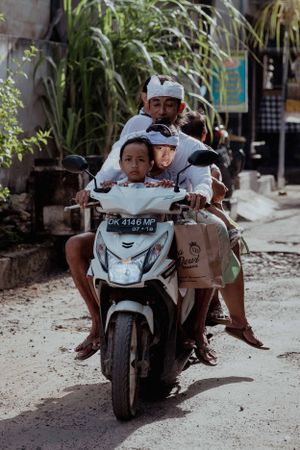 Riders on a motorbike traveling down a narrow street with lush vegetation.