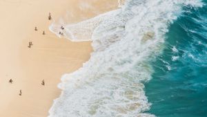 Aerial view of turquoise ocean waves meeting sandy beach with tiny beachgoers visible.