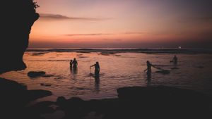 Silhouettes of beachgoers wading in shallow ocean water during dramatic sunset.