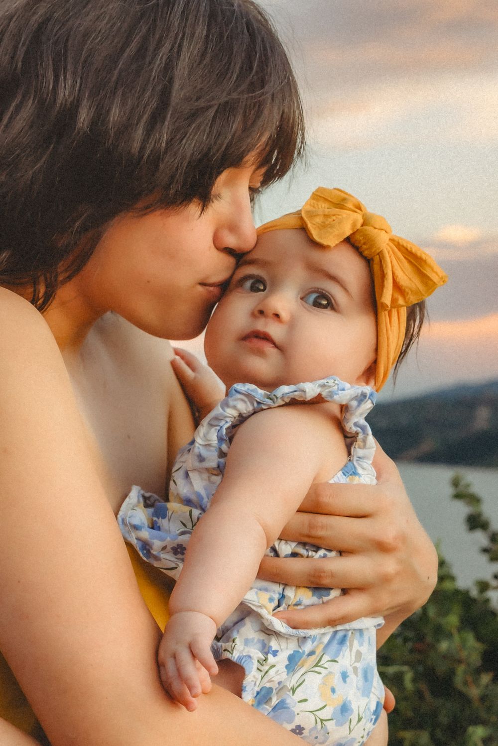 Tender moment between parent and baby in floral outfit with yellow bow at sunset against mountain backdrop.