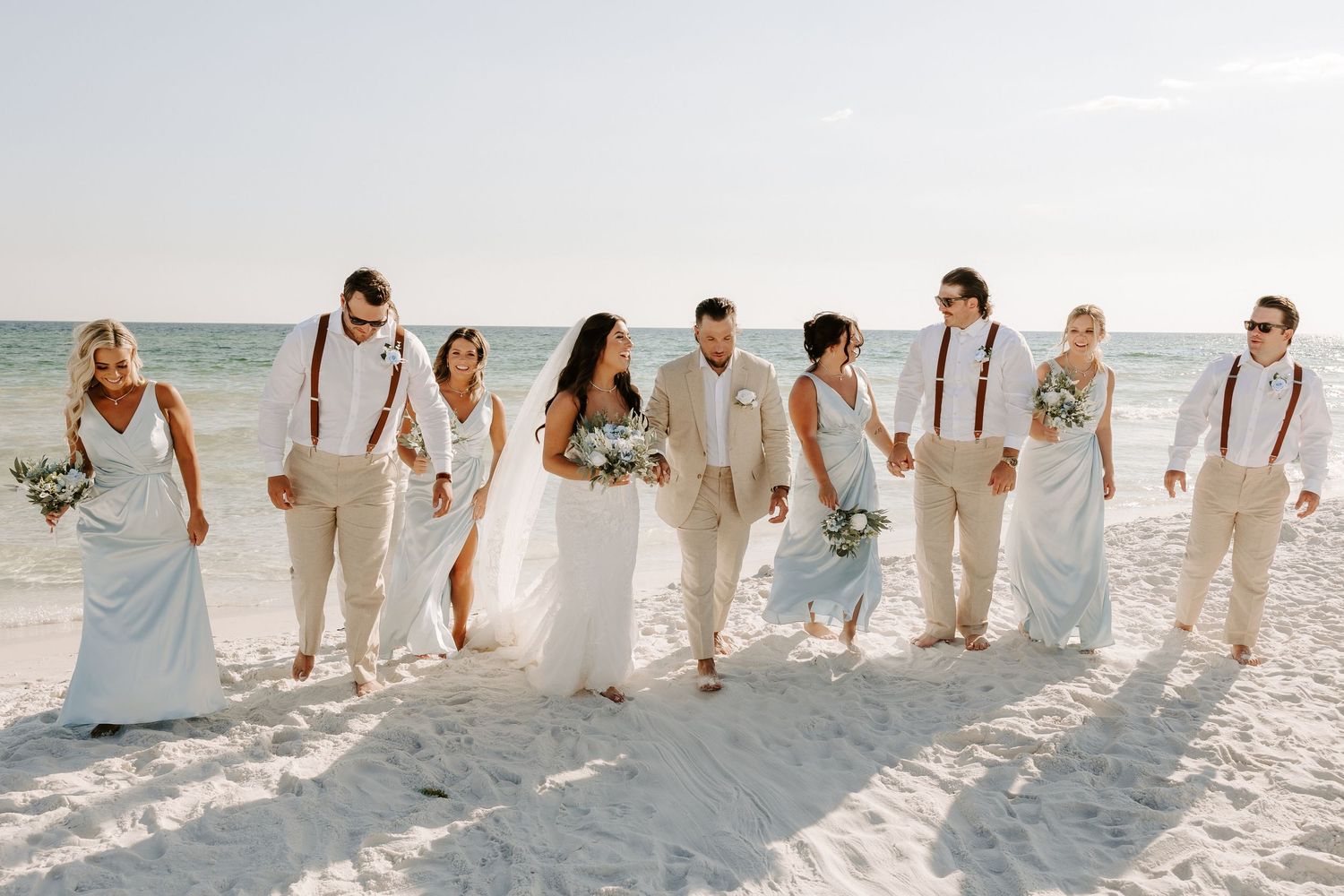 Wedding party walking together on sunny beach wearing white and beige outfits with suspenders.