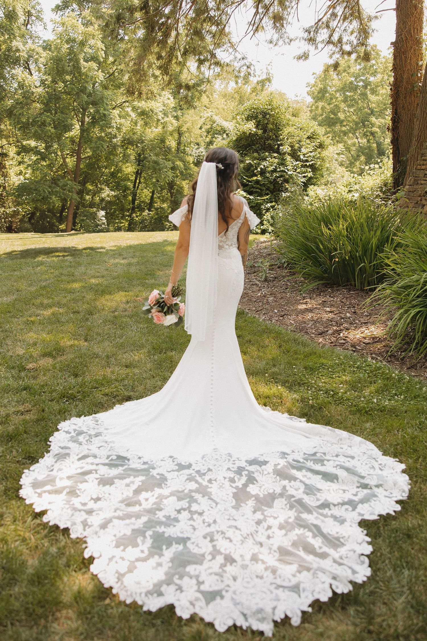 A stunning lace wedding gown with dramatic cathedral train photographed from behind in a sunlit garden setting.