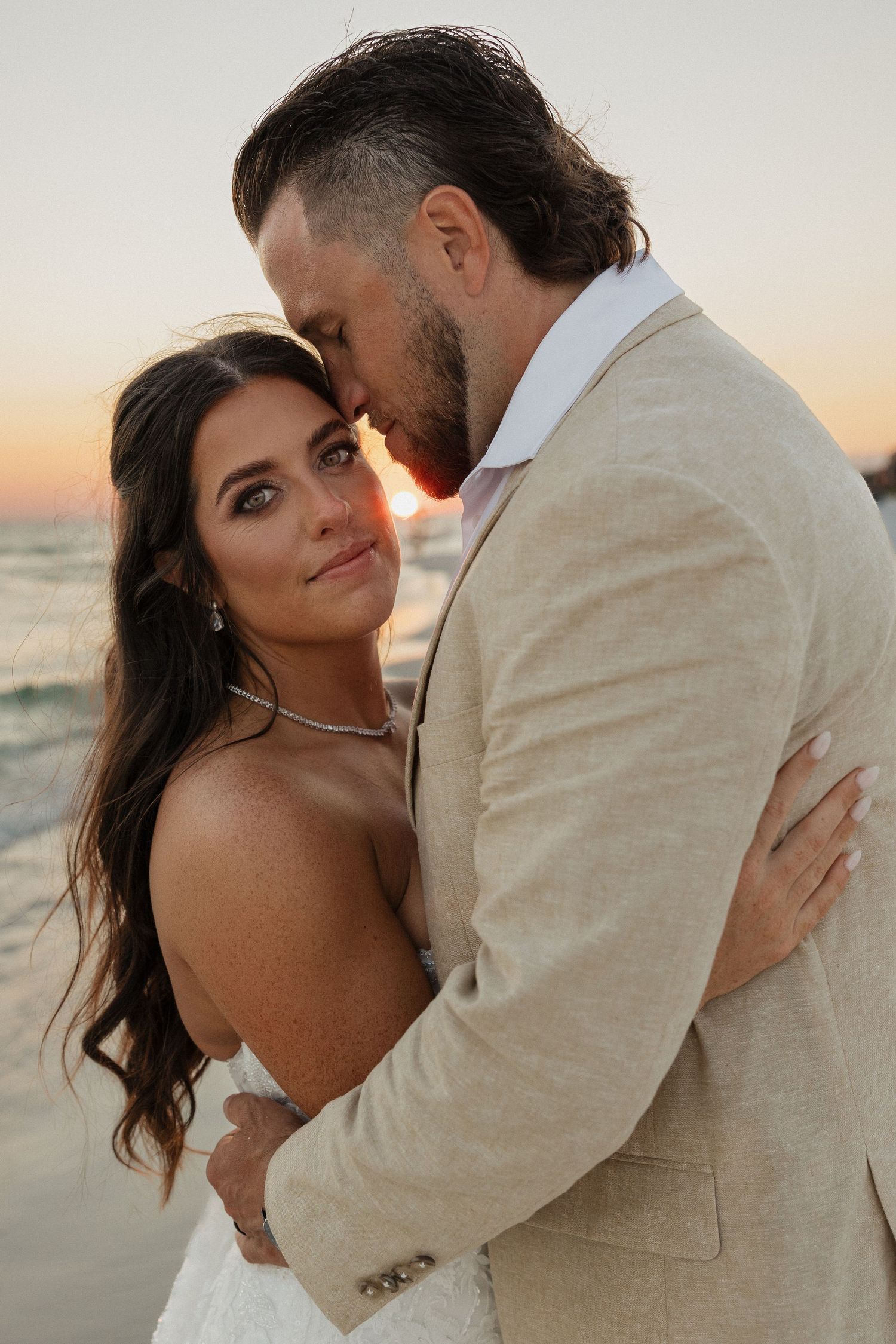 Romantic couple embraces on beach at sunset with warm lighting and beige formal attire.