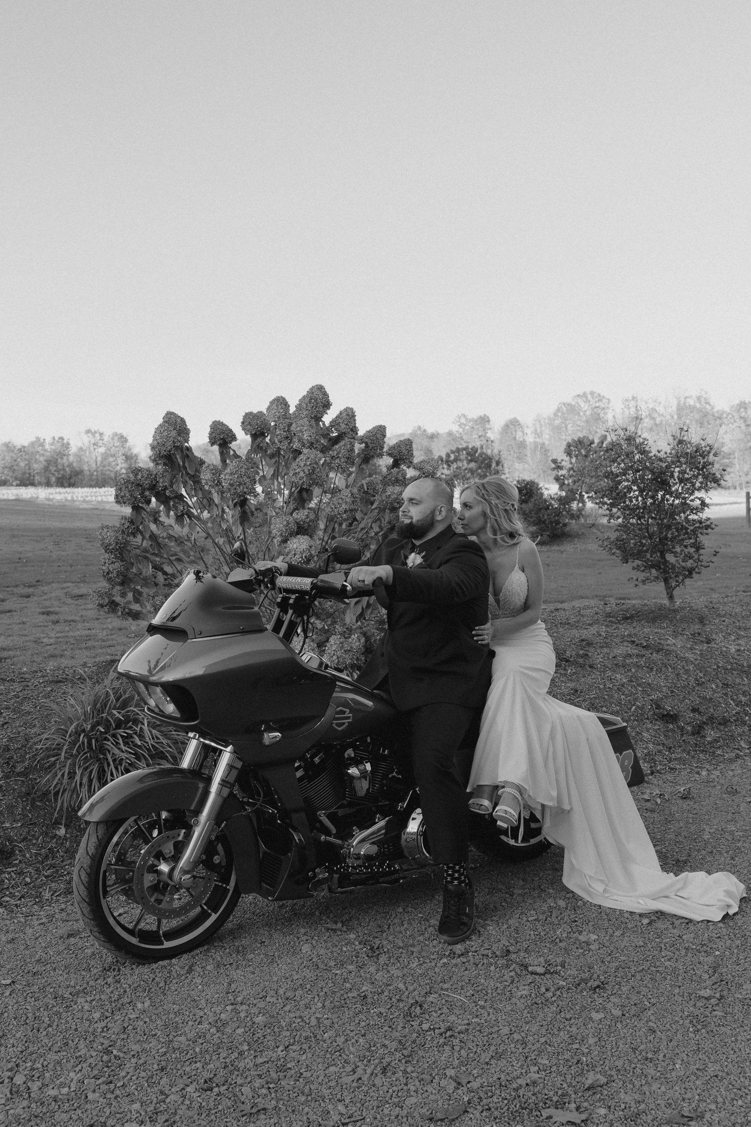 A black and white photo of newlyweds posing with a motorcycle in a rustic outdoor setting.