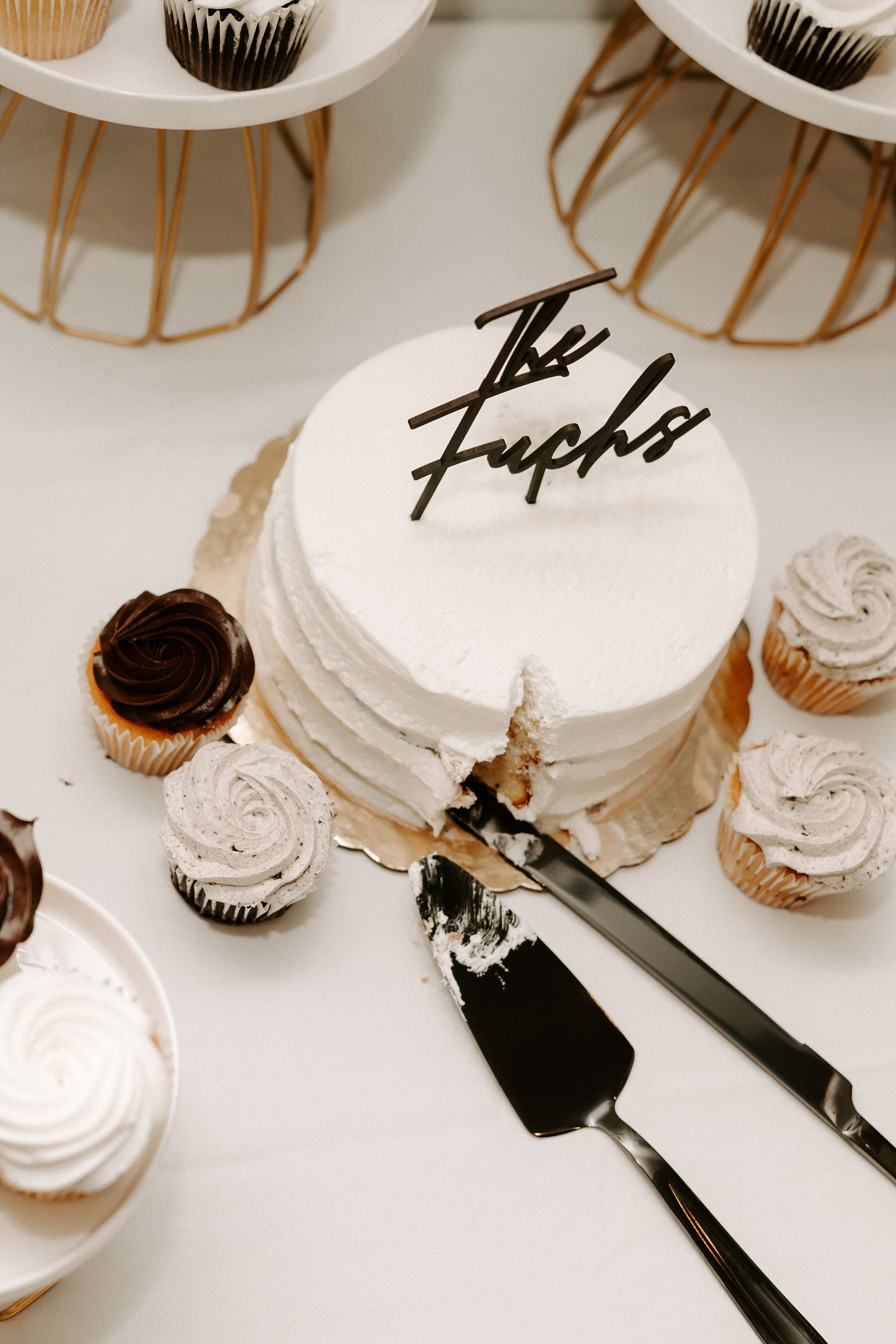 White wedding cake with black script lettering surrounded by chocolate and vanilla cupcakes.