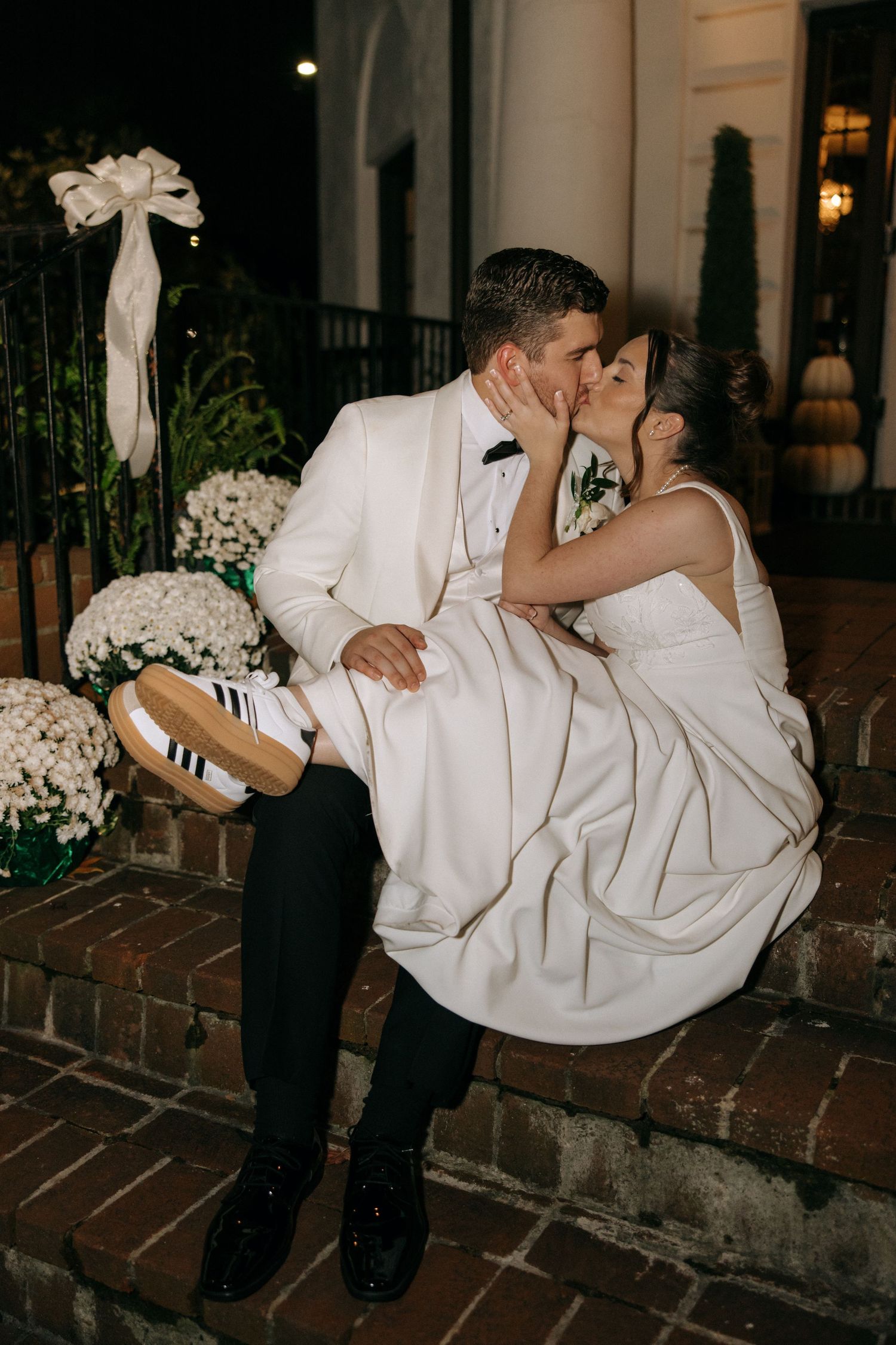 A romantic wedding couple shares a kiss on brick steps, with the bride wearing a white gown and striped sneakers.