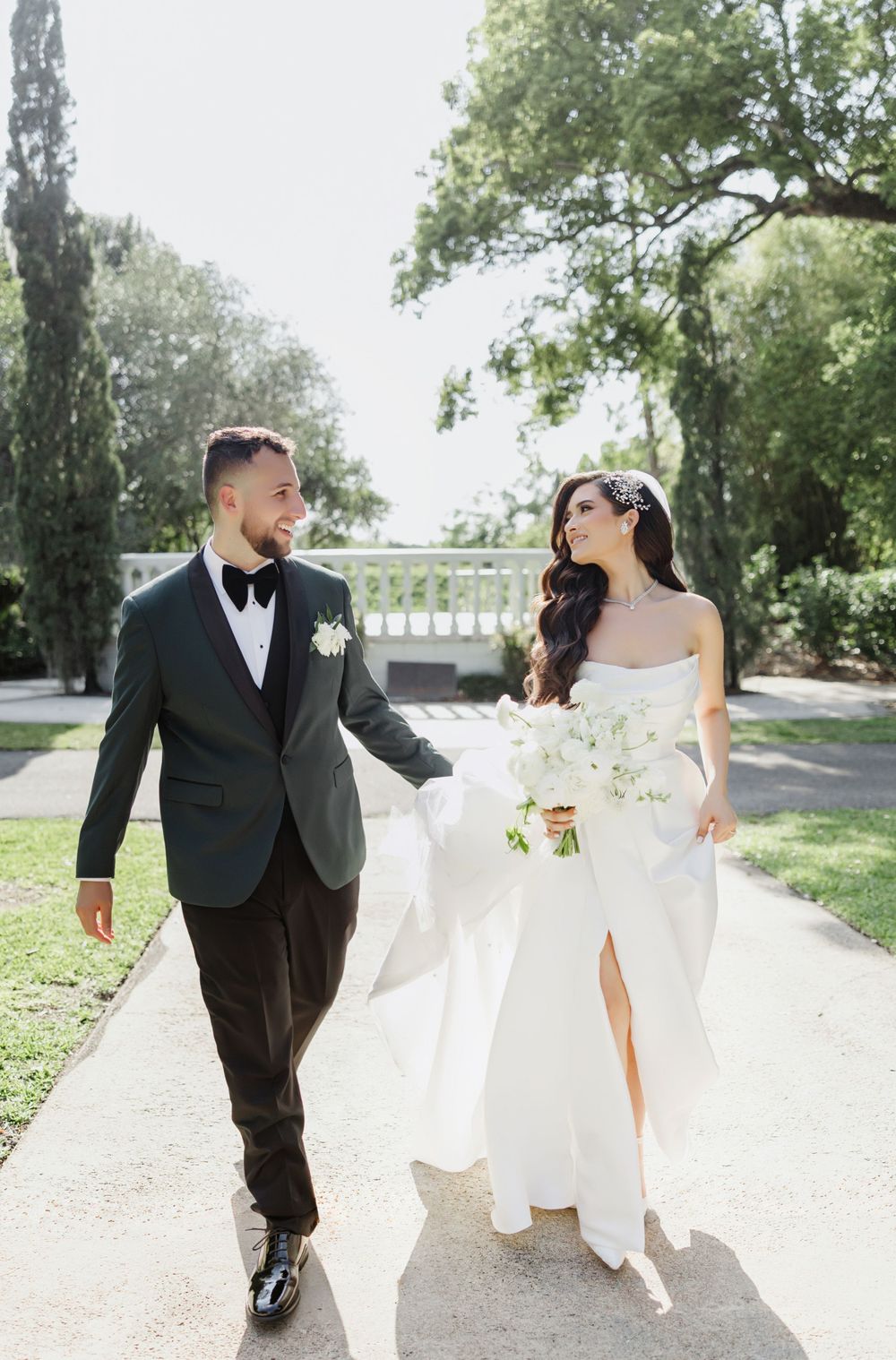 Bride and groom walk hand in hand along a sunlit path, smiling at each other in elegant wedding attire.