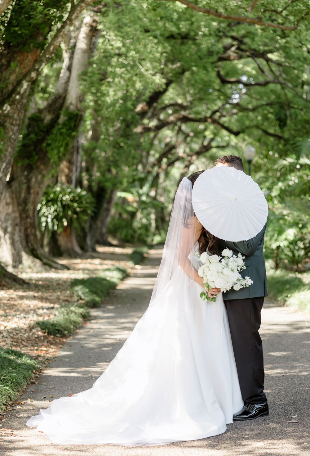 Bride and groom kissing on a tree-lined path, bride holding white bouquet and parasol in lush garden wedding setting.