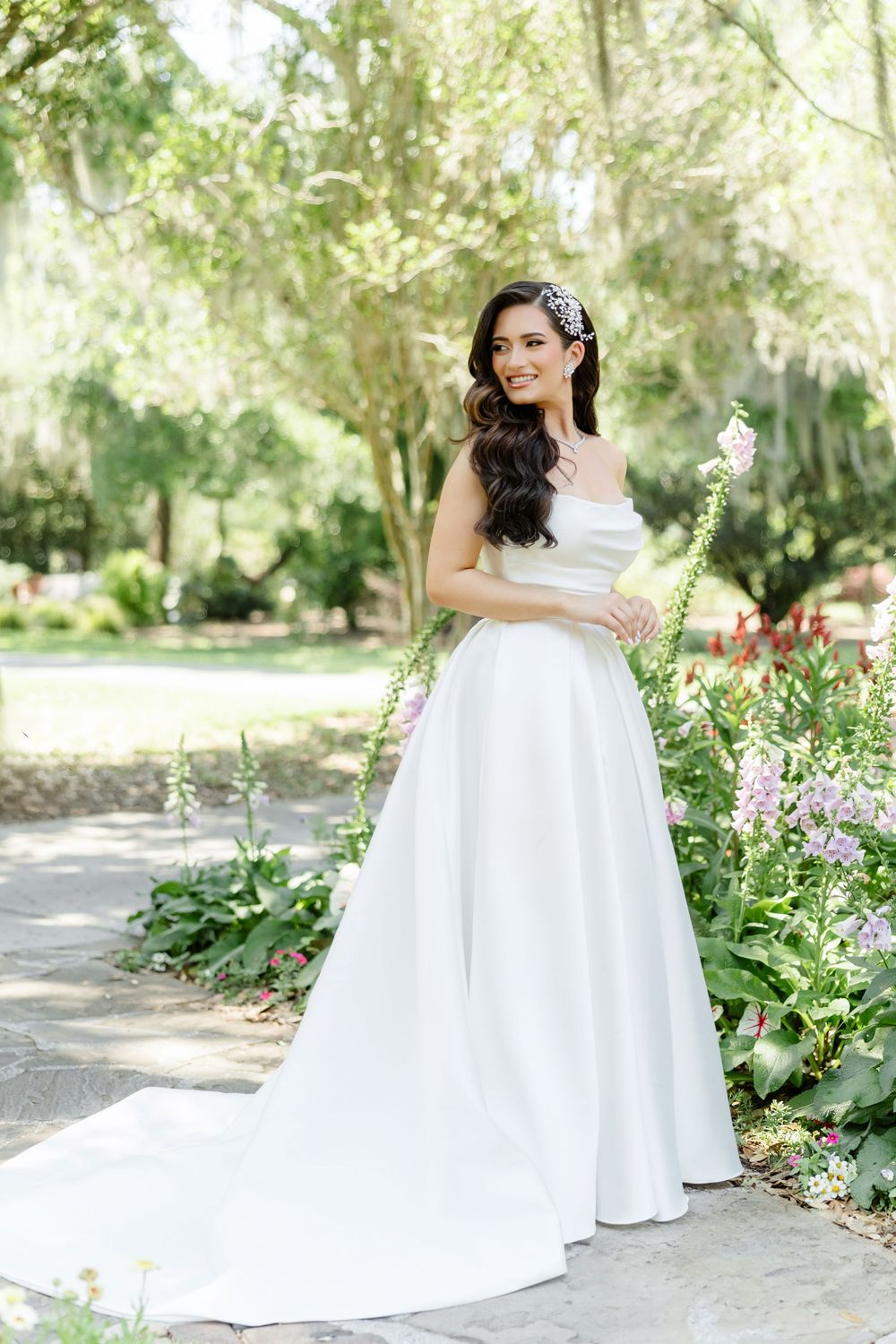 Smiling bride in elegant white off-shoulder ball gown with floral headpiece poses in lush garden setting.