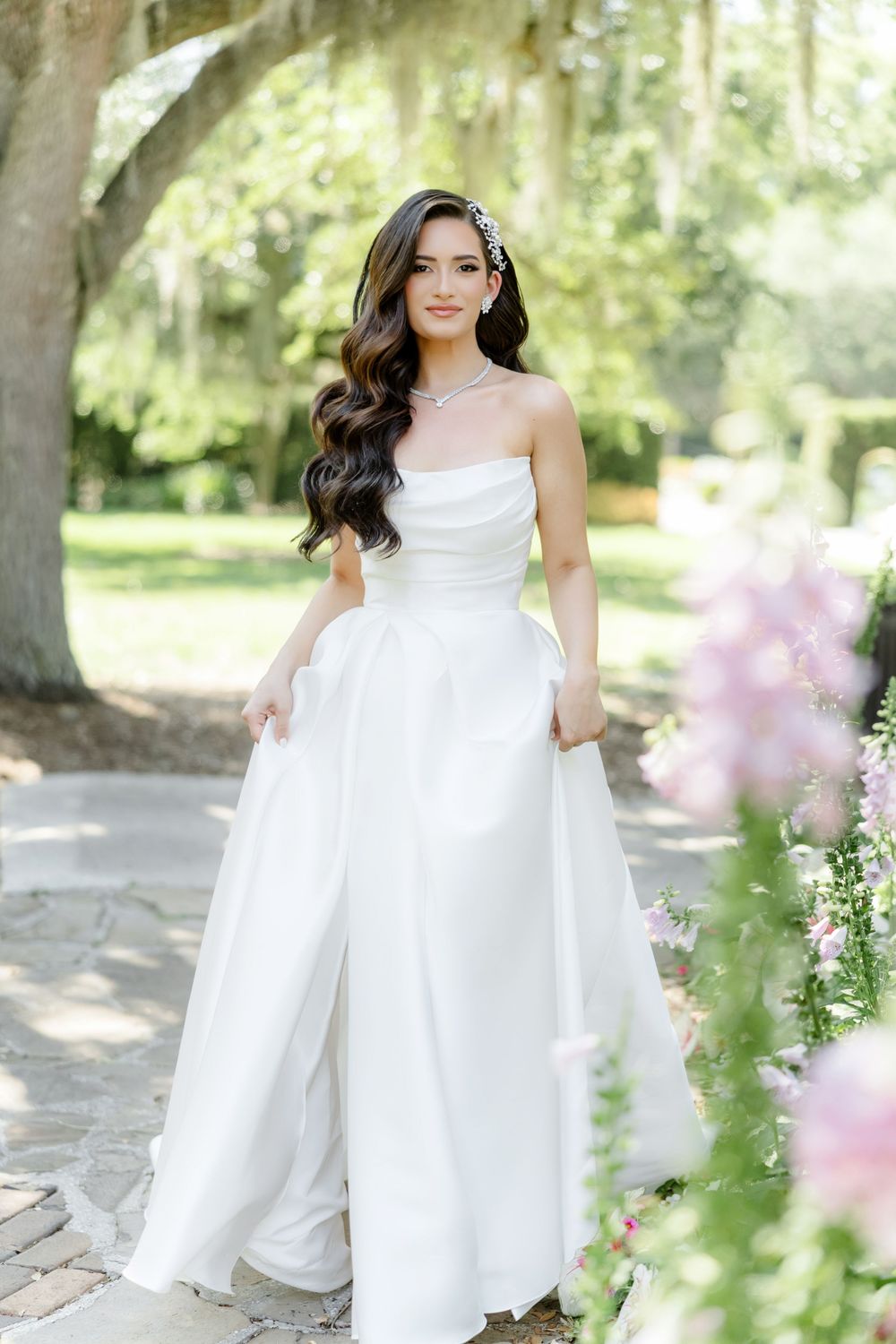 Bride in strapless white ball gown with pockets poses outdoors among pink flowers and moss-draped trees.