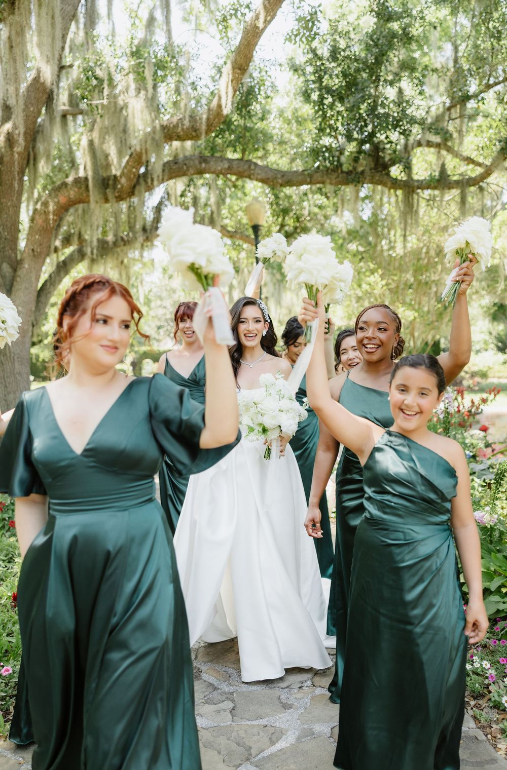 Bride in white gown walks with bridesmaids in emerald green satin dresses, raising bouquets outdoors.