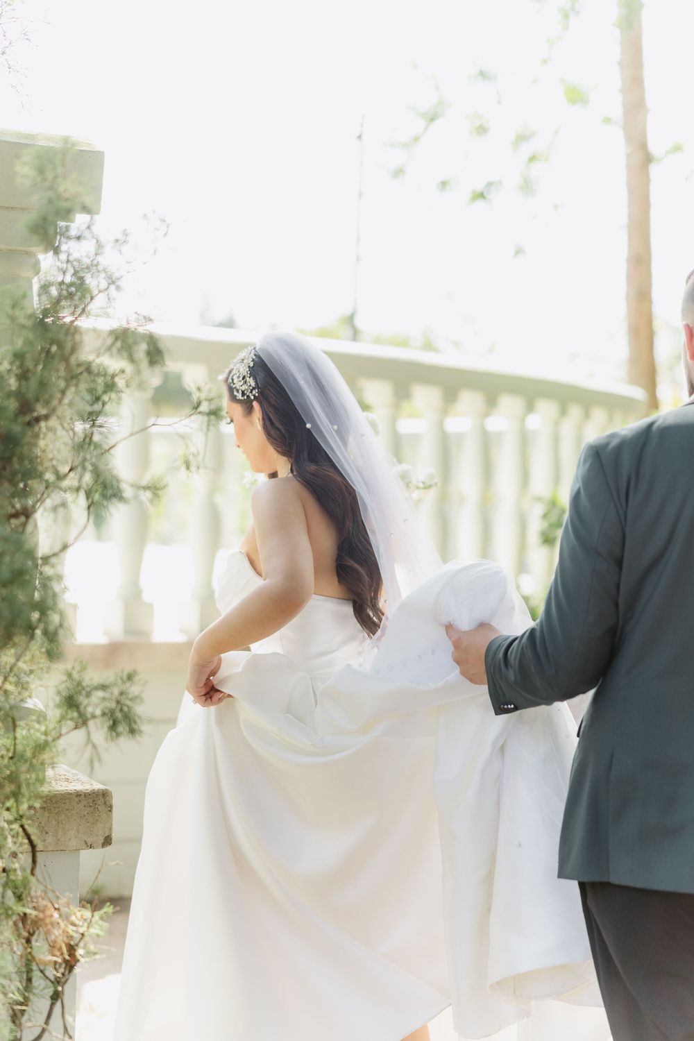 Bride in white gown and veil walks with groom in gray suit on elegant outdoor balcony surrounded by greenery.