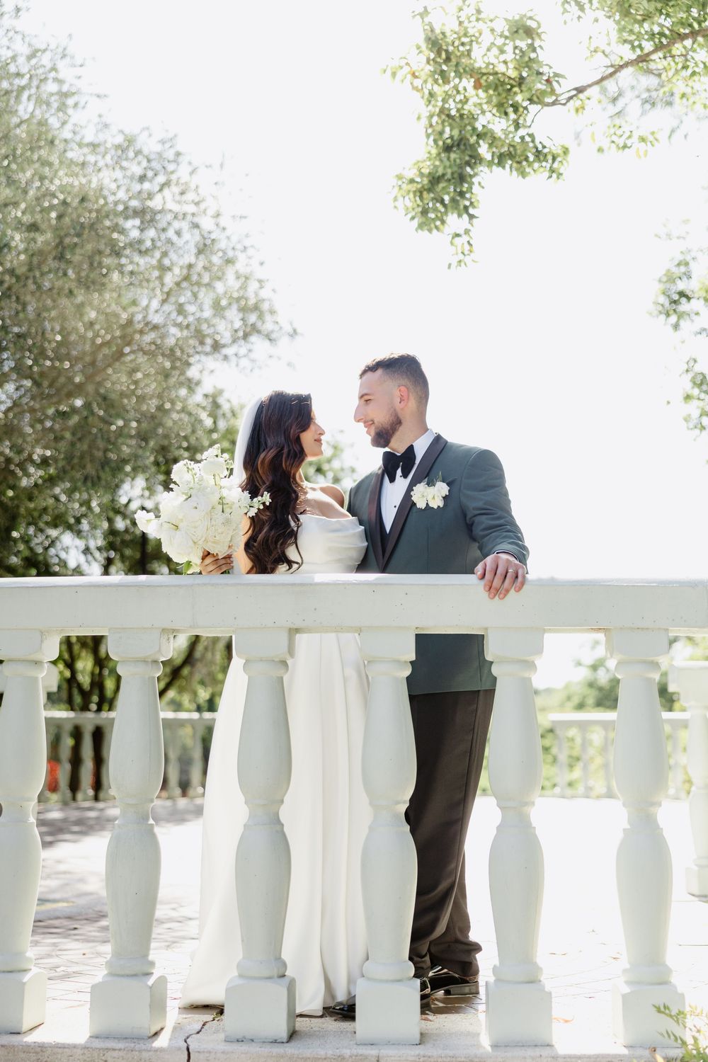 Bride and groom share a romantic moment on a white balustrade terrace, she holds white bouquet, he wears gray tuxedo.