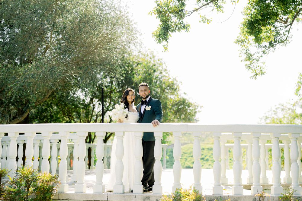 Elegant wedding couple poses on a classic white balcony railing in a garden setting.