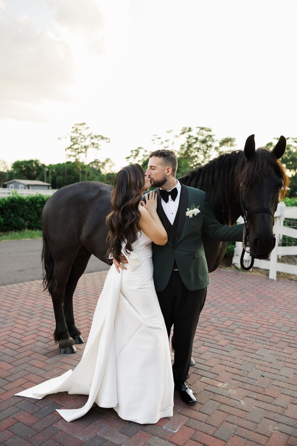 Wedding couple embraces on brick path with black horse standing beside them.